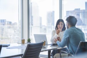 Business woman and man meeting and talking. Both are casually dressed. There is a laptop computer on the table. They are sitting at a boardroom table with a window behind them