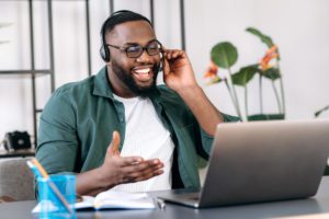 Friendly smiling African American guy, freelancer, student, confident call center operator in a headset, or ceo, uses laptop for video conference with coworkers or clients, telecommunication concept