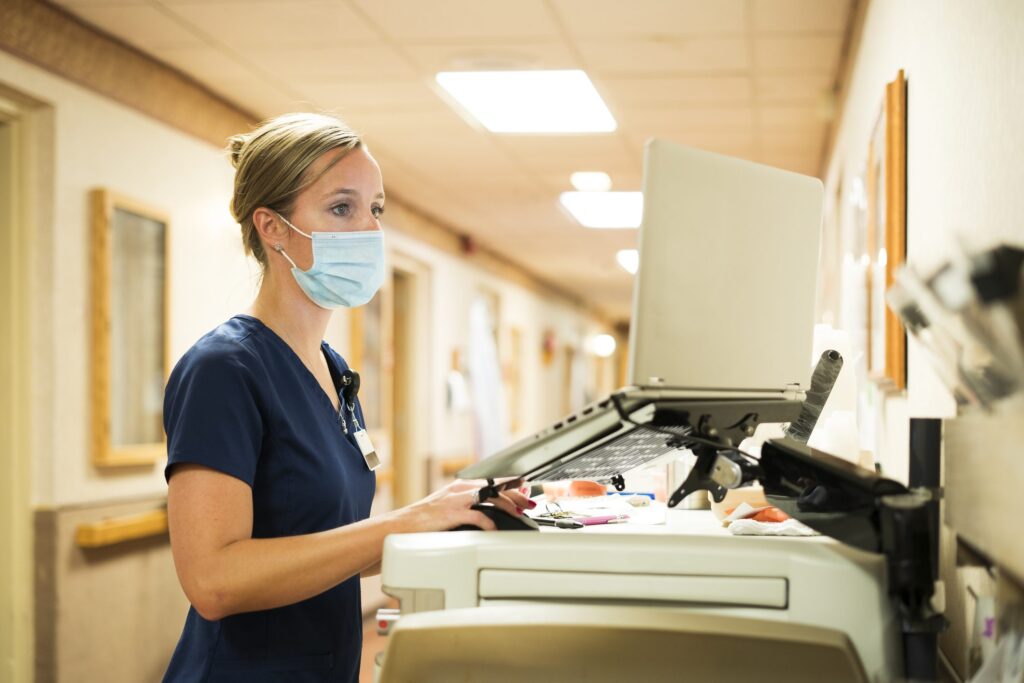 Registered nurse wearing a face mask documents medicine provided to a patient into a laptop computer at the nursing station at work, Indiana, USA