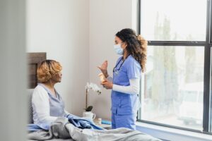 A female nurse gestures as she discusses a medication's side effects with a senior patient during a home visit. The patient is sitting in her bed.